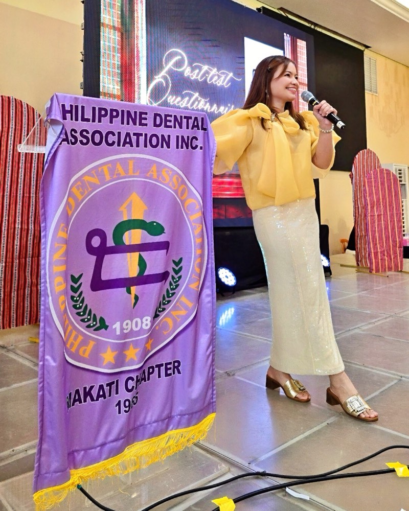 Dr. KC speaking into a microphone on stage beside a Philippine Dental Association Makati Chapter banner